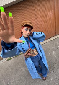 Casaco longo azul sobre uma blusa azul e calças largas azuis. Chapéu castanho, bolsa estampada. Unhas verdes brilhantes, maquiagem dramática nos olhos e tatuagem visível.
