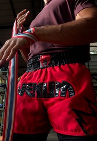 Crvene Venum kratke hlače s crnim elastičnim pojasom, sjajnim detaljima i teksturiranom površinom. Dizajn uključuje upečatljive logotipe i uzorke.