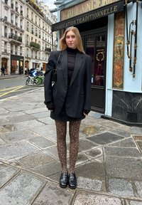 Zwarte oversized blazer over een fitted zwart top, korte zwarte rok en bruine luipaardprint leggings, aangevuld met zwarte loafers.