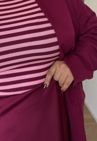 Magenta neuletakki vaaleanpunaisen ja burgundin raidallisen paidan päällä, jossa näkyy ribbineule. Sormi pitää kiinni sileästä, yksivärisestä magentasta kankaasta.