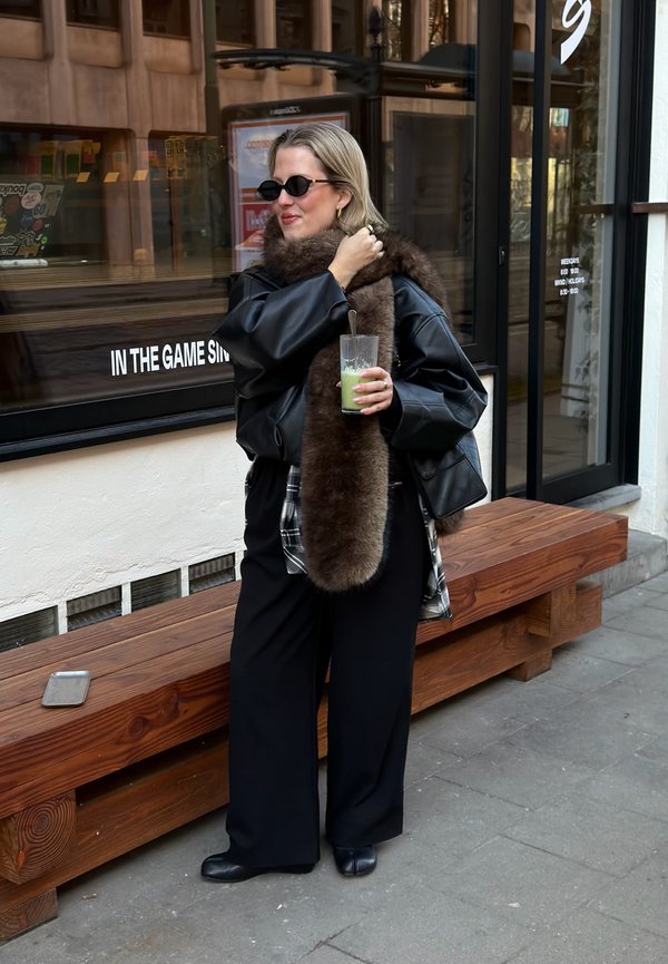 Femeie purtând o jachetă de piele neagră și un fular maro din blană, ținând un smoothie verde, stă lângă o bancă de lemn în fața unei cafenele.