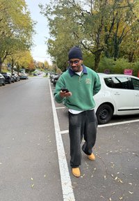 Grön sweatshirt med marinblå krage, svarta vida byxor och bruna stövlar, stående på en gata medan personen tittar på en smartphone.