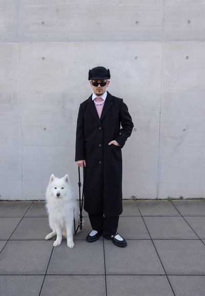 Un abrigo largo negro sobre un suéter rosa, gafas de sol y un sombrero negro; junto a un perro Samoyedo blanco con una correa, de pie sobre un pavimento gris.