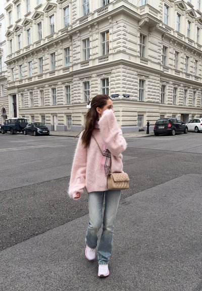 Joven mujer con un suéter rosa peludo y vaqueros caminando por una calle de la ciudad, sosteniendo la mano cerca de la cara, bolso beige sobre el hombro.