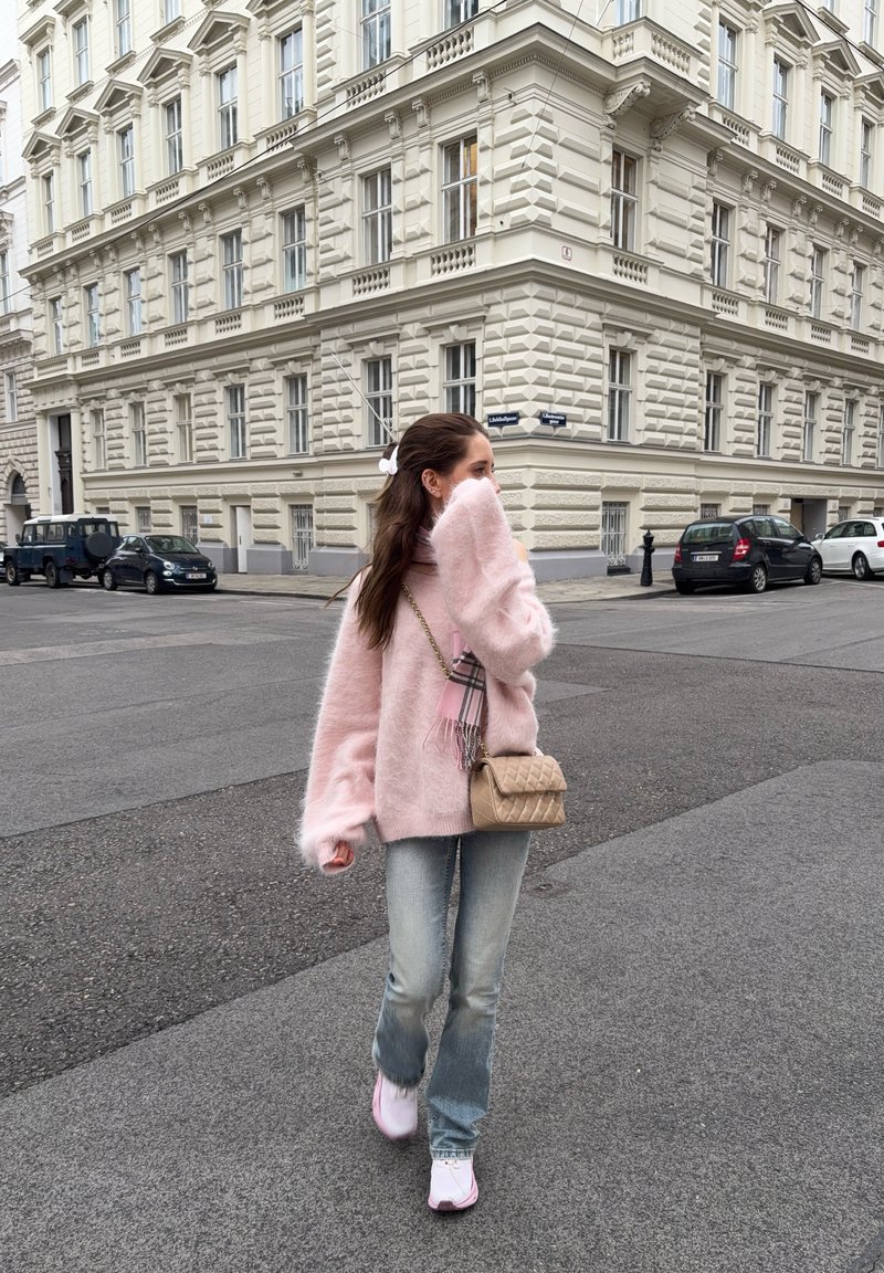 Jonge vrouw in een roze pluizige trui en jeans loopt op een stadstraat, houdt haar hand dicht bij het gezicht, draagt een beige handtas over de schouder.