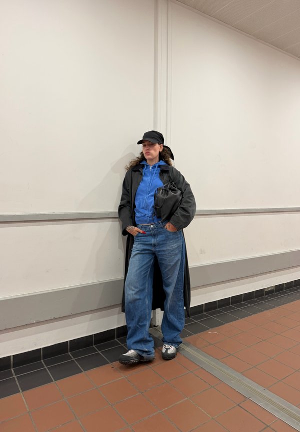 Frau mit schwarzer Mütze, langem schwarzem Mantel, blauem Kapuzenpullover und weitem blauen Jeans steht an einer weißen Wand auf einem Fliesenboden und hält eine schwarze Tasche.