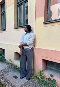 Sivý pletený sveter cez košeľu s golierom, skombinovaný s voľnými sivými nohavicami. Čierne topánky, stojaci pred pastelovou stenou.