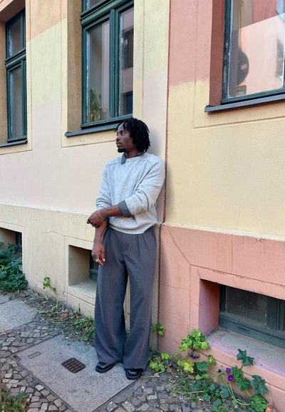 Suéter gris texturizado sobre una camisa con cuello, combinado con pantalones grises sueltos. Zapatos negros, de pie contra una pared pastel.