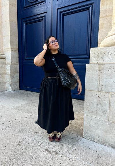 Top corto negro, falda larga negra, bolso negro con detalles de cordones, zapatillas púrpuras y doradas. Puertas azules y pared de piedra en el fondo.