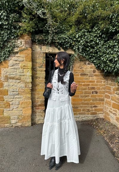 Mujer con vestido blanco de volantes y chaleco de encaje, cuello alto negro y botas, de pie junto a una pared de piedra cubierta de hiedra y una puerta arqueada.