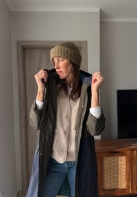 Femme portant un bonnet en laine et des manteaux superposés, ajustant le col de sa veste, debout à l'intérieur près d'une porte en bois et d'un meuble TV.