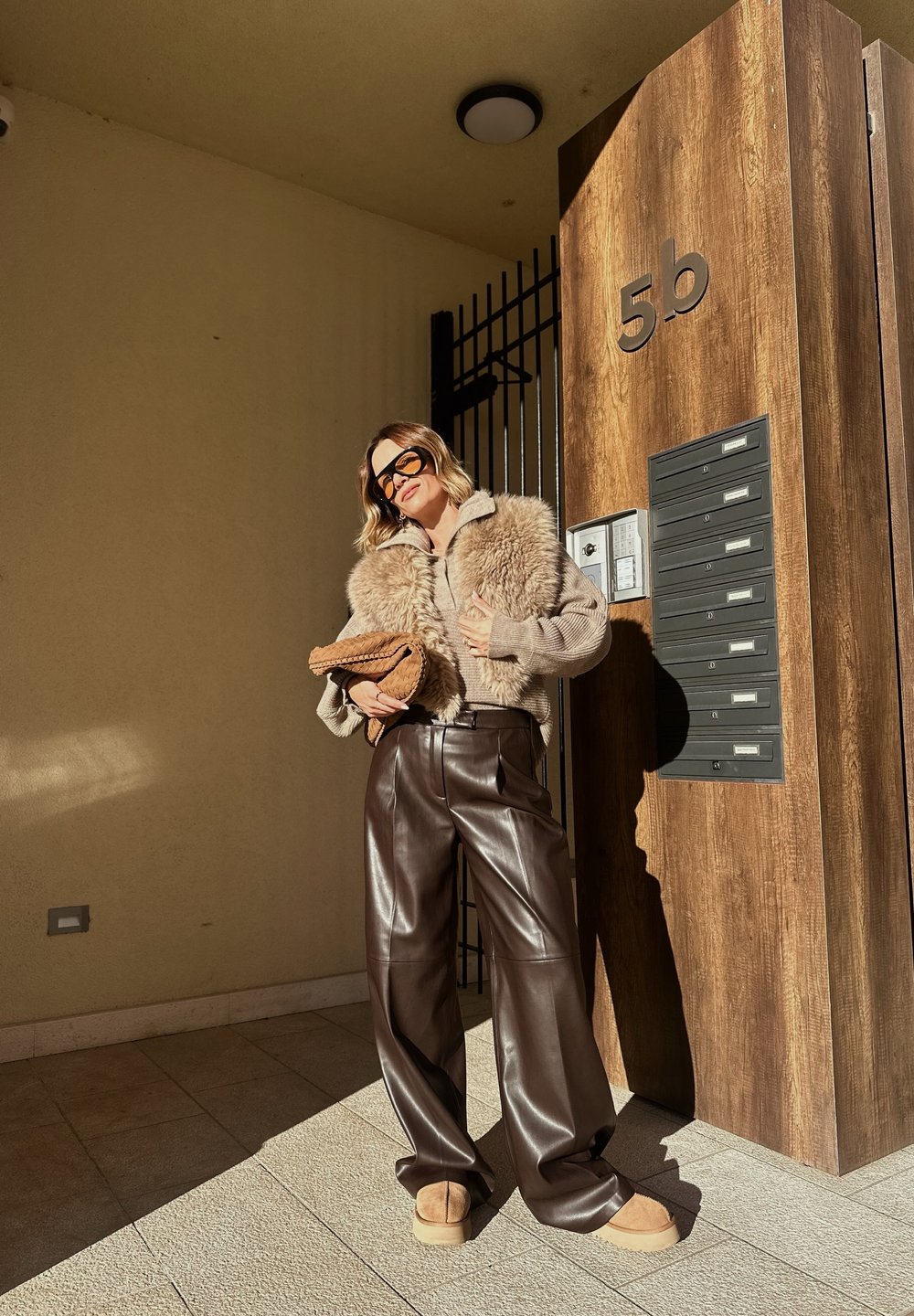 Femme portant des lunettes de soleil, un gilet en fourrure beige, un pull en tricot, un pantalon en cuir marron et des chaussures beige, se tenant près d'une porte en bois portant l'étiquette 5b et des boîtes aux lettres.