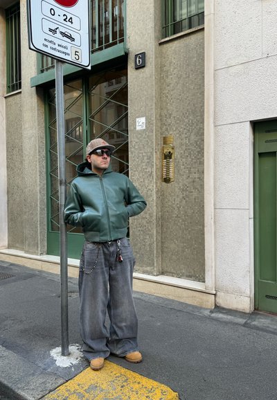 Chaqueta verde de cuero con cremallera, jeans grises de pierna ancha, botas marrones y una gorra estampada. De pie junto a un letrero de calle y una fachada de edificio texturizada.