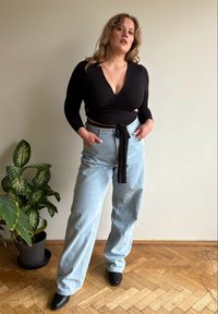 Juodas apvyniojamas viršus su ilgomis rankovėmis ir rišimo detale; šviesiai mėlynos spalvos, aukštu liemeniu džinsai; juodi kampuoti aulinukai; vidaus aplinka su augalu.