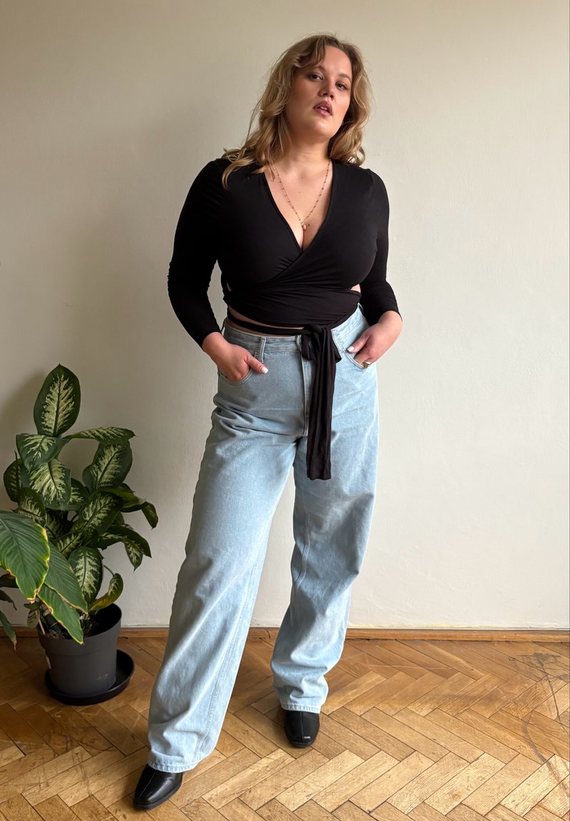 Musta kietaisupaita pitkillä hihoilla ja solmittavalla yksityiskohdalla; vaaleansiniset korkeavyötäröiset farkut; mustat teräväkärkiset nilkkurit; sisätiloissa, viereen sijoitettu kasvi.