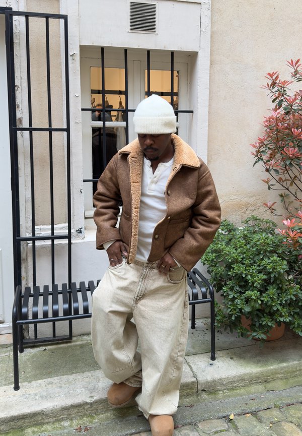 Mann trägt eine braune Lammfelljacke, weiße Strickmütze, übergroße beige Hose und beige Schuhe und steht in der Nähe einer schwarzen Bank und Blumentöpfe.