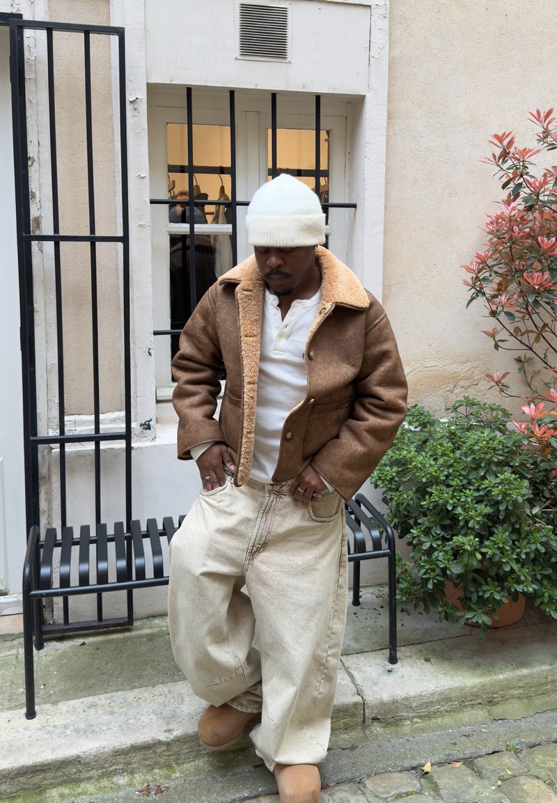 Άνδρας που φορά καφέ δερμάτινο μπουφάν με επένδυση γούνας, λευκό πλεκτό σκουφάκι, φαρδιά μπεζ παντελόνια και καφέ παπούτσια στέκεται κοντά σε ένα μαύρο παγκάκι και φυτά σε γλάστρες.