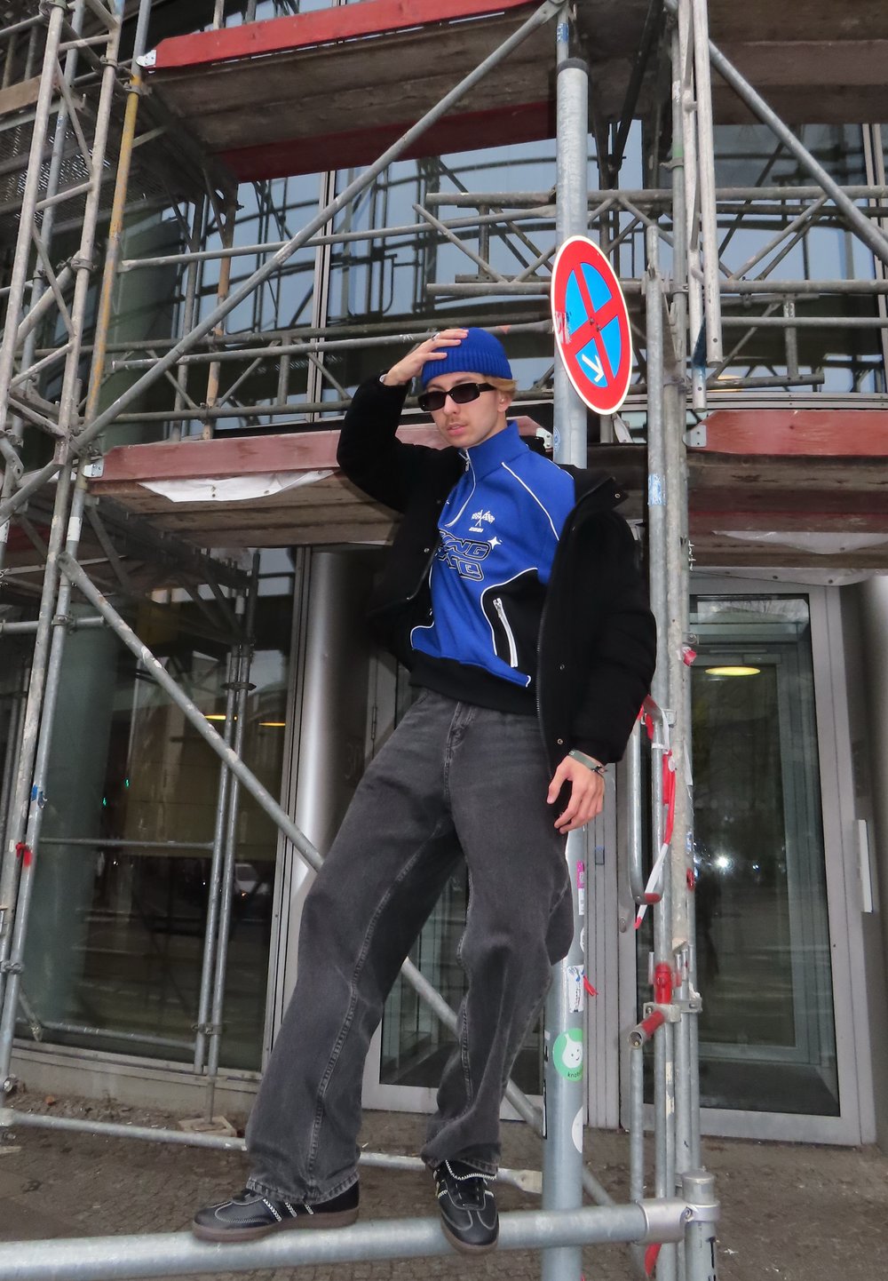 Blå glidelåsjakke over svart skjorte, grå vide bukser, svarte sko, blå lue og solbriller; pose på et stillas med et parkeringsforbudsskilt.