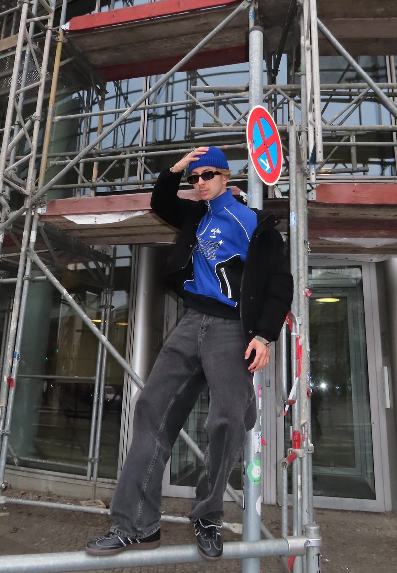 Giacca blu con zip sopra maglietta nera, pantaloni grigi a gamba larga, scarpe nere, berretto blu e occhiali da sole; posato su impalcature con un segnale di divieto di sosta.