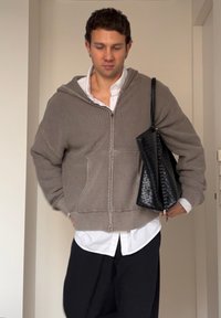 Lichtbruine geribde hoodie met ritssluiting, gedragen over een wit shirt. Zwarte textuur tas op de schouder. Loszittende zwarte broek. Eenvoudig, casual ontwerp.