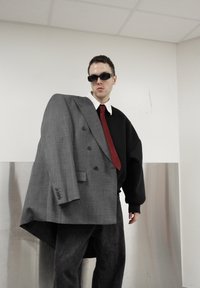 Blazer gris surdimensionné à double boutonnage avec un sweat-shirt noir asymétrique. Une cravate rouge, un pantalon sombre et des lunettes de soleil complètent le look.