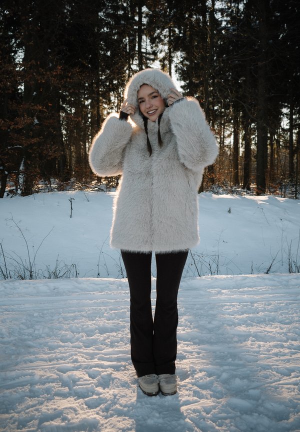 Junge Frau in weißem Fellmantel und schwarzer Hose lächelt im Freien auf verschneitem Boden mit Wald im Hintergrund bei Sonnenuntergang.