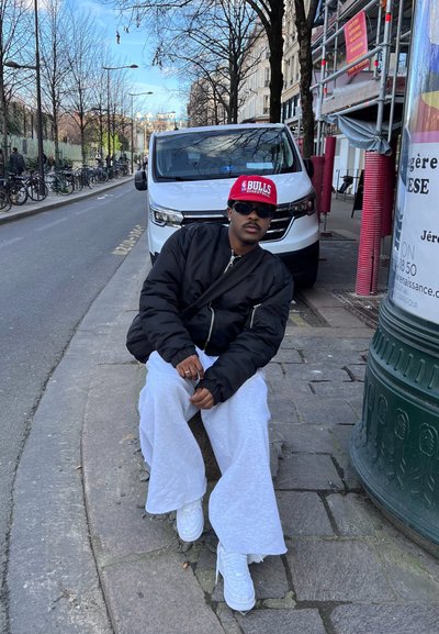 Chaqueta bomber negra, gorra roja "BULLS", pantalones anchos blancos. Sentado en la acera cerca de una furgoneta blanca y un objeto urbano verde. Cielo despejado de fondo.