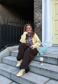 Jeune femme portant des lunettes, une écharpe colorée, un pull jaune et un pantalon marron, assise sur des marches en pierre avec une tasse de café et un sac à main bleu clair.