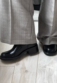 Botas de tornozelo pretas com acabamento em couro liso e salto robusto, combinadas com calças largas cinza com padrão xadrez. Fundo de piso de madeira.
