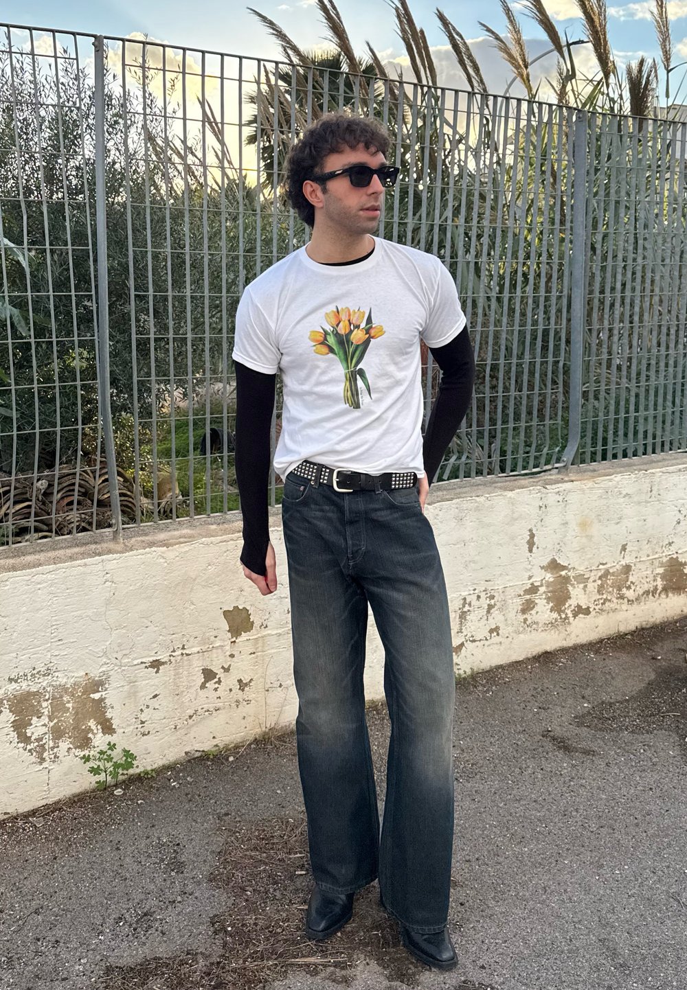 Λευκό μπλουζάκι με γραφικό μπουκέτο κίτρινων τουλιπών, επικαλυμμένο με μαύρα μακριά μανίκια. Τζιν σε στυλ καμπάνας και μαύρες μπότες, μπροστά από φράχτη.