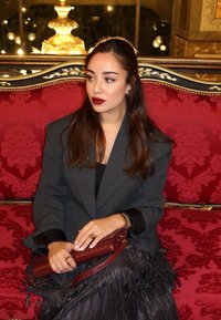 Donkergrijs colbert over een zwart shirt, een rode leren handtas vasthoudend. Lang, golvend bruin haar; donkerrode lippen; zittend op een weelderige rood-patroon bekleding.