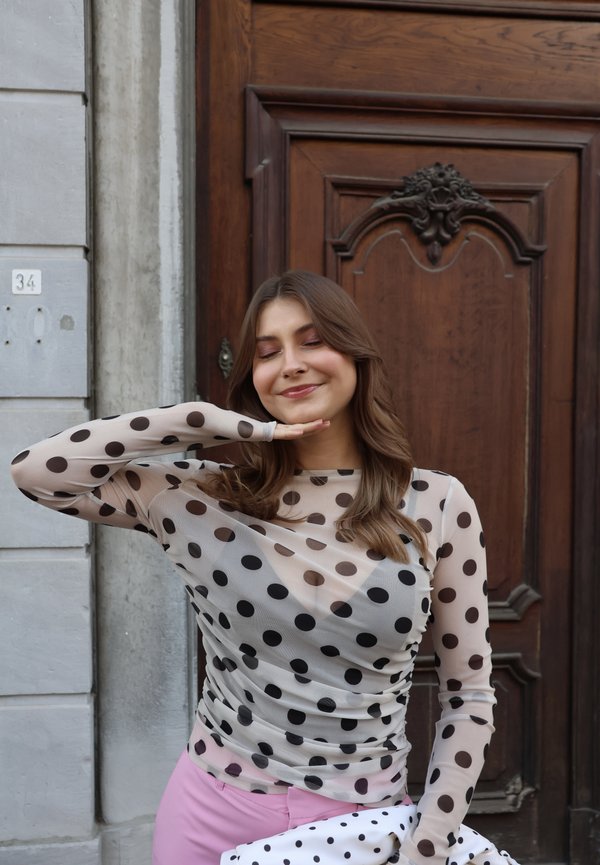 Jonge vrouw in doorschijnende top met stippen en roze broek poseert met gesloten ogen, hand onder de kin, staand bij een houten deur en stenen muur.