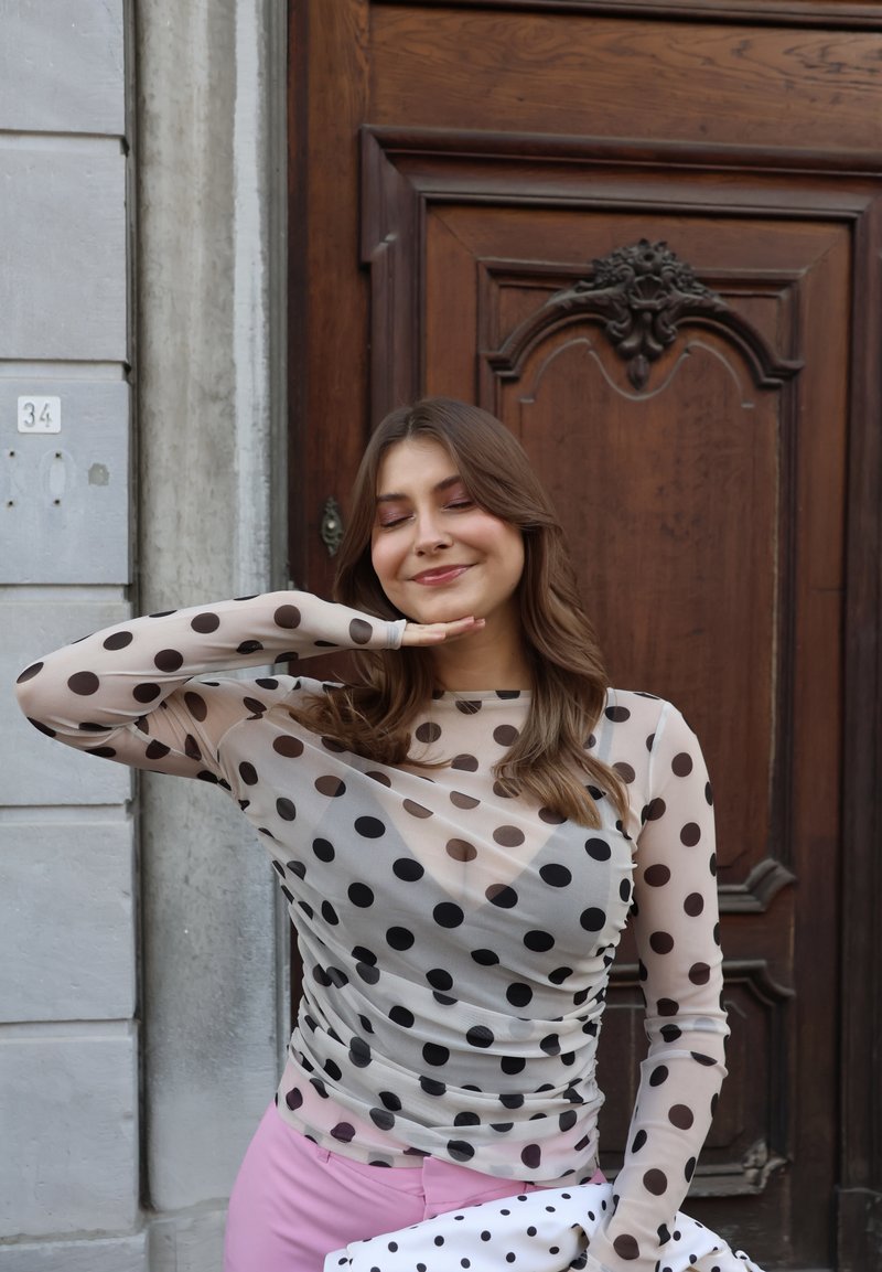 Jeune femme vêtue d'un haut transparent à pois et d'un pantalon rose, posant les yeux fermés, la main sous le menton, debout près d'une porte en bois et d'un mur en pierre.