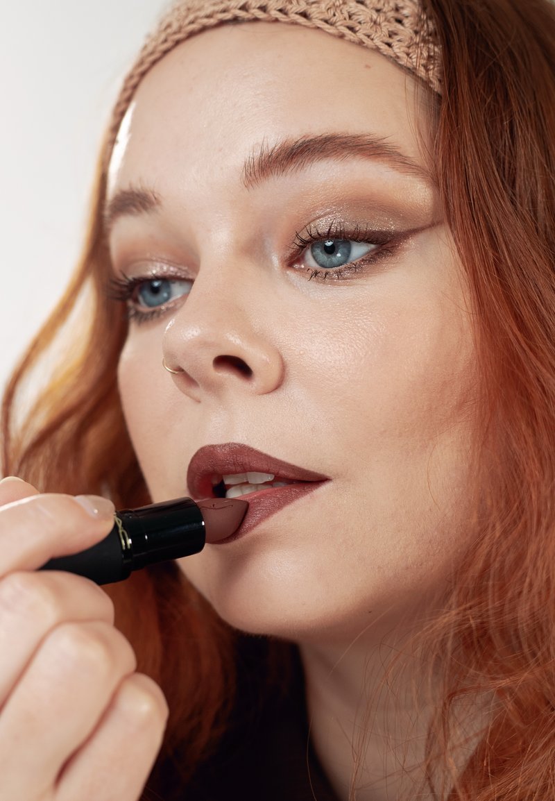 Femme aux cheveux roux et aux yeux bleus appliquant un rouge à lèvres mauve foncé, portant un bandeau en tricot beige et un maquillage des yeux subtil.