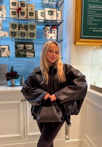 Czarna skórzana, oversize'owa kurtka z bufkami na rękawach, zestawiona z małą torebką. W tle półki z różnymi produktami Ralph Lauren.