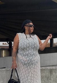 Balta nėrinių suknelė su laisva forma, turinti gėlių raštą. Modelis dėvi akinius nuo saulės ir juodą kepurę, laiko juodą rankinę.