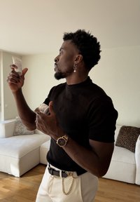 Homme en chemise noire et pantalon blanc lisant les étiquettes de deux contenants de soins de la peau dans un salon avec des canapés blancs.