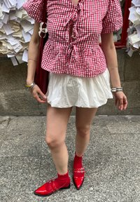 Czerwono-biała krata top peplum z bufkami, biała plisowana mini spódniczka oraz błyszczące czerwone czółenka zakończone szpicem z klamrami i czerwonymi skarpetkami.