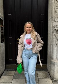 Beżowy płaszcz typu trench na białej T-shircie z krótkim rękawem i grafiką czerwonej truskawki, jasnoniebieskie dżinsy z wysokim stanem oraz zielona pikowana torba.
