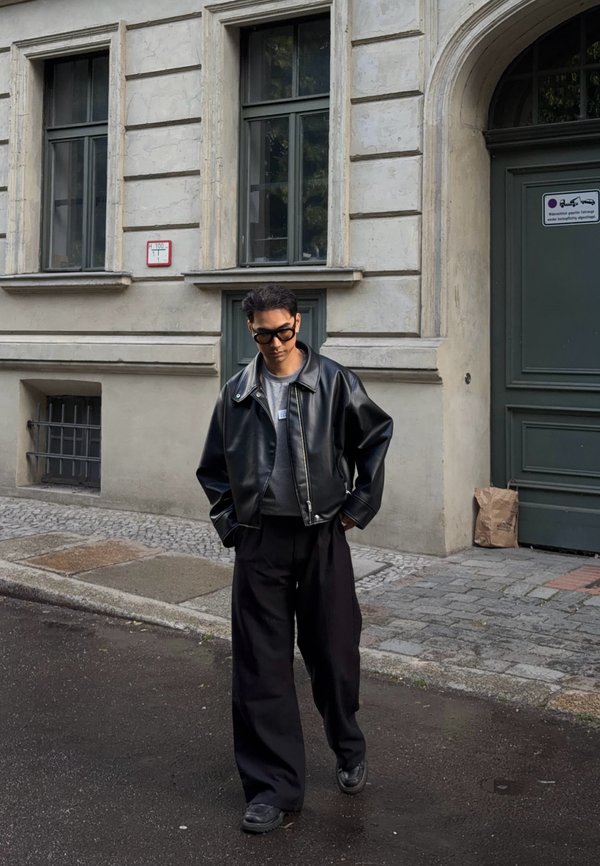 Jeune personne portant une veste en cuir noire, un pantalon large et des lunettes de soleil foncées marchant dans une rue mouillée devant un bâtiment beige avec de grandes fenêtres.