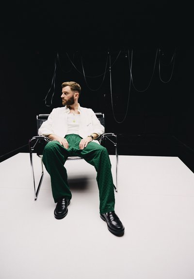 Sillas e individuo se encuentran contra un fondo oscuro. El individuo lleva una chaqueta blanca, pantalones verdes texturizados y zapatos negros. Silla moderna de cromo.