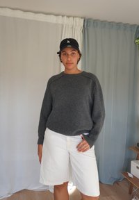 Siv pleteni pulover z raglan rokavi, v kombinaciji z belimi šortami. Model nosi črno kapo in obroče za ušesa. Ozadje vsebuje svetle zavese.