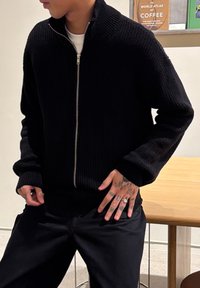 Άτομο που φοράει μαύρο ζακέτα με φερμουάρ και μαύρο παντελόνι, με χέρια καλυμμένα με τατουάζ να ακουμπούν στους γοφούς, στέκεται κοντά σε ένα ξύλινο τραπέζι και βιβλία.