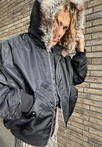 Sort bomberjakke med pelsforet hætte, med lynlås foran, elastiske manchetter og løs pasform. Tekstureret stof med glans.