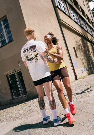 Camiseta Asics GEL-Kayano en blanco con estampado gráfico, combinada con pantalones cortos negros. El modelo tiene tatuajes y lleva zapatillas en azul claro y rojo.