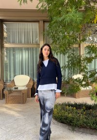 Jeune femme aux longs cheveux foncés, portant un pull marine et une jupe à motifs, debout sur une terrasse près de chaises en osier et de verdure.