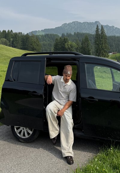 Vehículo negro con la puerta lateral abierta, que presenta a una persona con una camisa a rayas y pantalones beige, sentada en el borde. Fondo de paisaje verde.