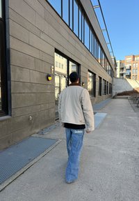 Lys beige jakke med brun krage, svart skjorte og brede blå jeans. Innstillingen har store vinduer og teksturerte grå vegger.