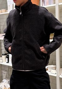 Personne portant une veste zippée noire texturée, les mains dans les poches, debout dans un magasin avec des étagères de céramiques et de produits en arrière-plan.