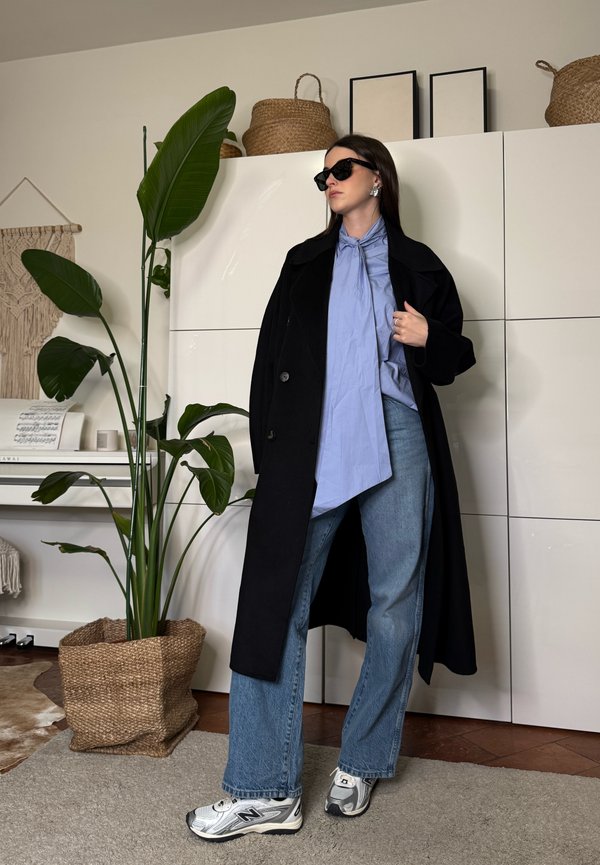 Femeie purtând o haină neagră, bluză albastră cu cravată, blugi largi și adidași, stând lângă o plantă mare în ghiveci într-o cameră modernă.